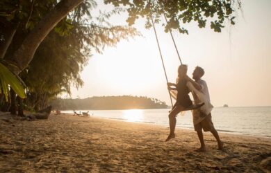 A couple enjoys a beach swing at sunset. The man gently pushes the woman, both smiling, surrounded by lush trees and calm waves. The golden sky reflects on the water, creating a serene and romantic atmosphere.