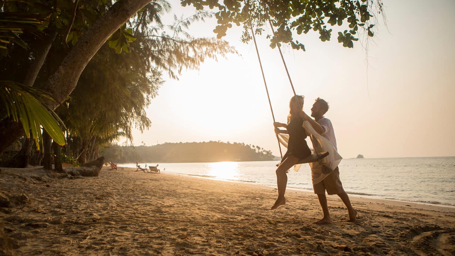 A couple enjoys a beach swing at sunset. The man gently pushes the woman, both smiling, surrounded by lush trees and calm waves. The golden sky reflects on the water, creating a serene and romantic atmosphere.