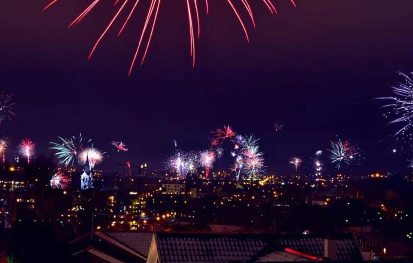 Fireworks light up the night sky above a city, illuminating the landscape with bursts of colorful lights. Buildings and rooftops are faintly visible beneath the vibrant display.