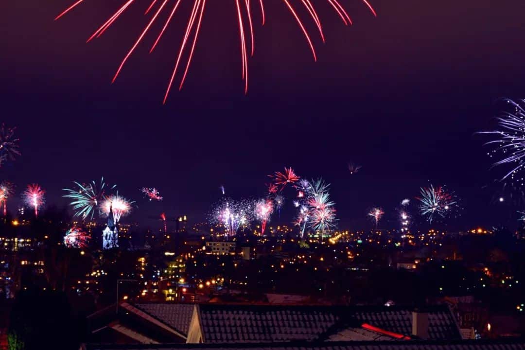 Fireworks light up the night sky above a city, illuminating the landscape with bursts of colorful lights. Buildings and rooftops are faintly visible beneath the vibrant display.