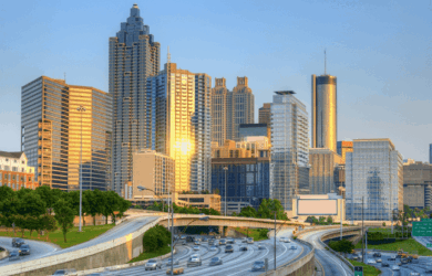Skyline of a bustling city at sunset, featuring tall, modern skyscrapers with reflective glass. A multi-lane highway curves through the foreground, filled with moving vehicles. The sky is clear, casting a warm glow over the buildings.