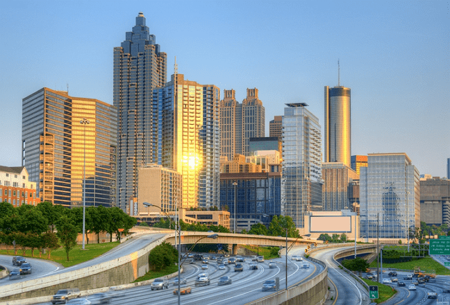 Skyline of a bustling city at sunset, featuring tall, modern skyscrapers with reflective glass. A multi-lane highway curves through the foreground, filled with moving vehicles. The sky is clear, casting a warm glow over the buildings.