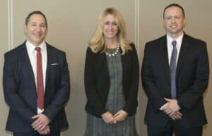 Three people in formal attire stand side by side, smiling. The person on the left wears a dark suit with a red tie, the person in the center wears a dark blazer over a patterned dress, and the person on the right wears a dark suit with a patterned tie.