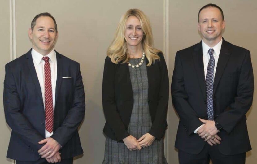 Three people in formal attire stand side by side, smiling. The person on the left wears a dark suit with a red tie, the person in the center wears a dark blazer over a patterned dress, and the person on the right wears a dark suit with a patterned tie.