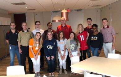 A group of thirteen people standing indoors, some wearing various college football jerseys. They are posing and smiling in a room with tables and chairs.