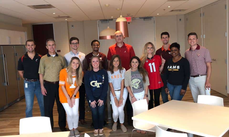 A group of thirteen people standing indoors, some wearing various college football jerseys. They are posing and smiling in a room with tables and chairs.