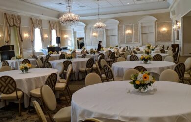 A large, elegant banquet hall with round tables covered in white tablecloths. Each table features a floral centerpiece. Chandeliers hang from the ceiling, and the room is decorated in neutral tones. A person is seen setting up the space.