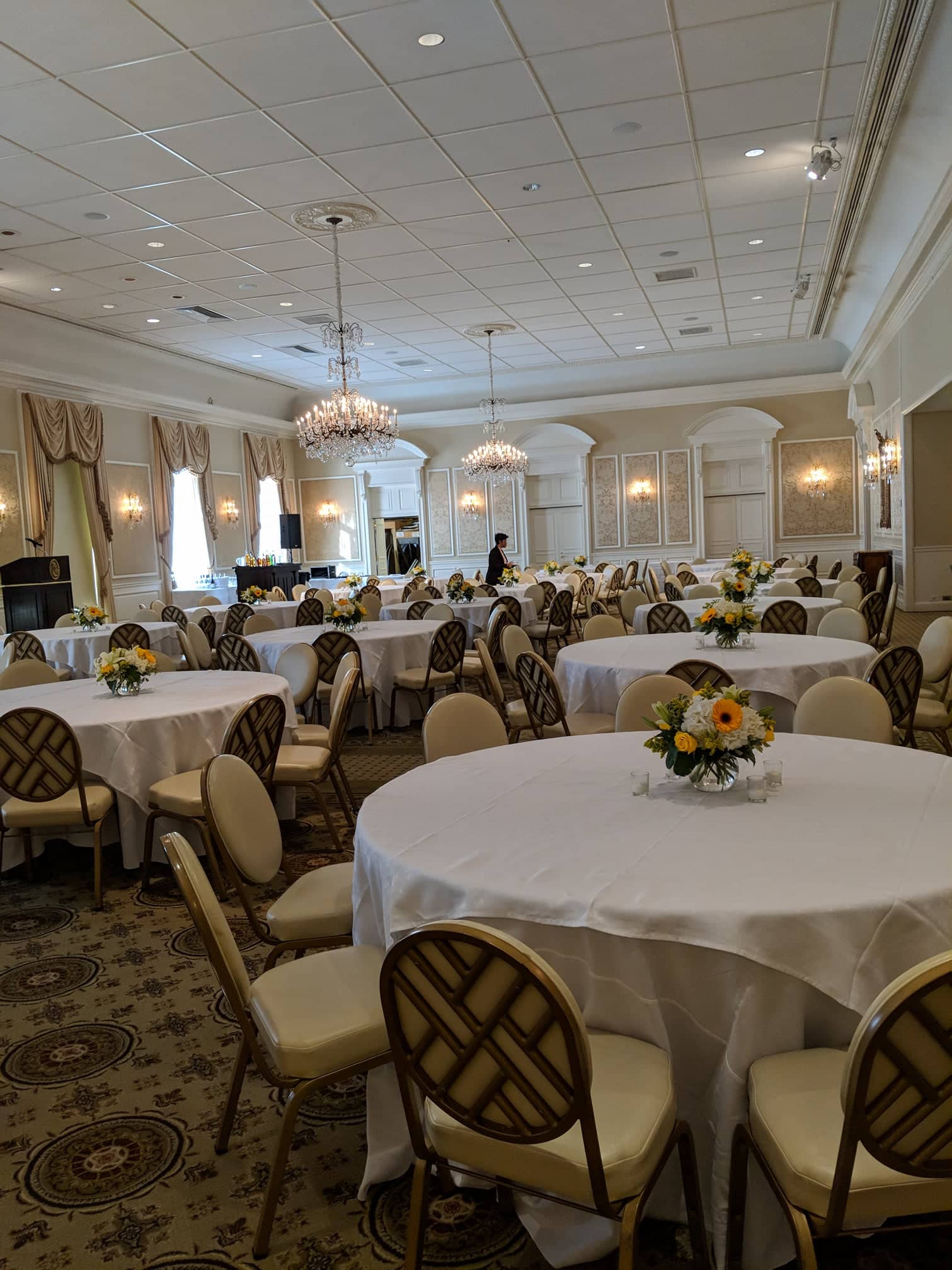 A large, elegant banquet hall with round tables covered in white tablecloths. Each table features a floral centerpiece. Chandeliers hang from the ceiling, and the room is decorated in neutral tones. A person is seen setting up the space.