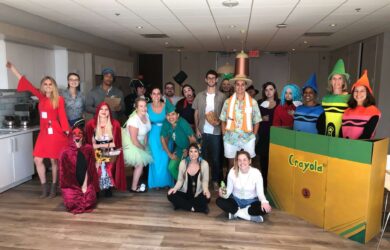 A group of people dressed in various colorful costumes, including crayon outfits, a devil, and fairy tale characters, pose together in a bright room with wooden flooring. A large Crayola crayon box prop is also visible.