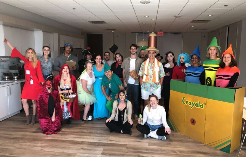 A group of people dressed in various colorful costumes, including crayon outfits, a devil, and fairy tale characters, pose together in a bright room with wooden flooring. A large Crayola crayon box prop is also visible.