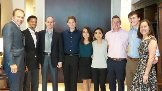 A group of nine people smiling and standing in a line in front of dark double doors. They are dressed in business casual attire, with some wearing shirts and others in blouses.