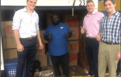 Four people stand in front of an open truck filled with cardboard boxes. Three men are giving thumbs up, while one stands inside the truck smiling. They appear to be in a loading area or warehouse.