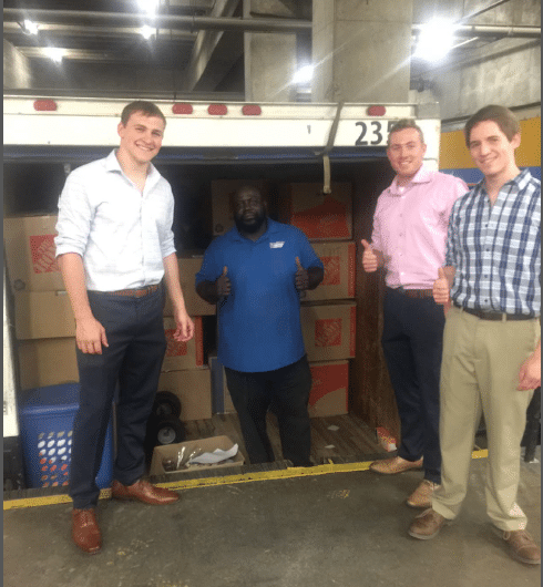 Four people stand in front of an open truck filled with cardboard boxes. Three men are giving thumbs up, while one stands inside the truck smiling. They appear to be in a loading area or warehouse.