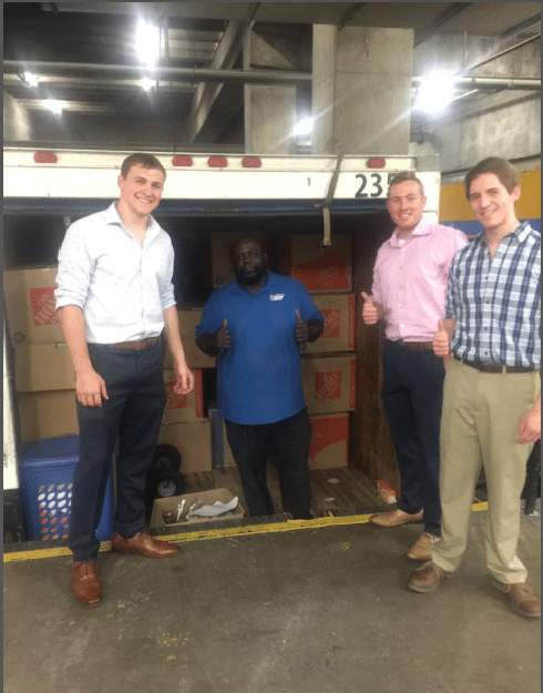 Four people stand in front of an open truck filled with cardboard boxes. Three men are giving thumbs up, while one stands inside the truck smiling. They appear to be in a loading area or warehouse.