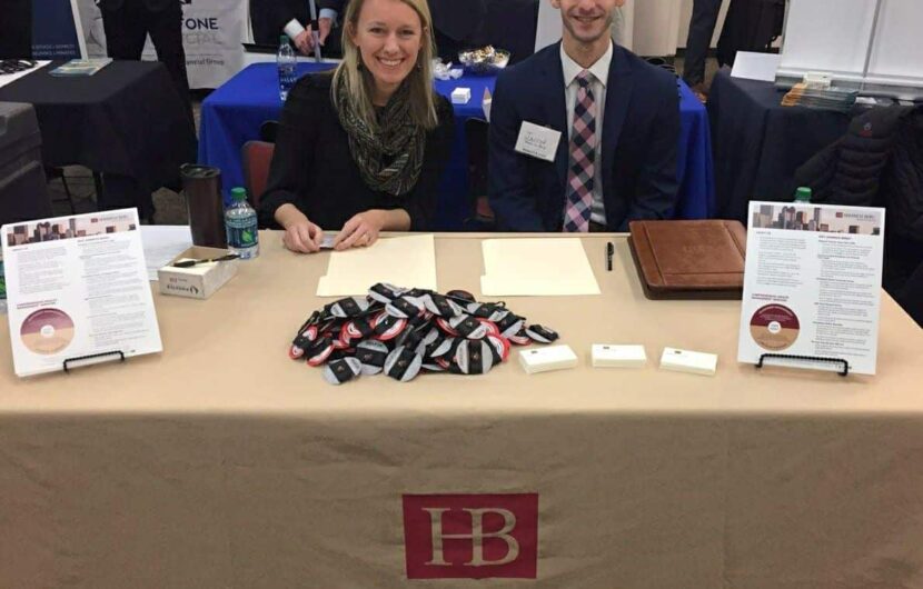 Two individuals sitting at a table with a beige cloth labeled Homrich Berg. The table has papers, a water bottle, name tags, and informational pamphlets. They are smiling and appear to be at a professional event or job fair.