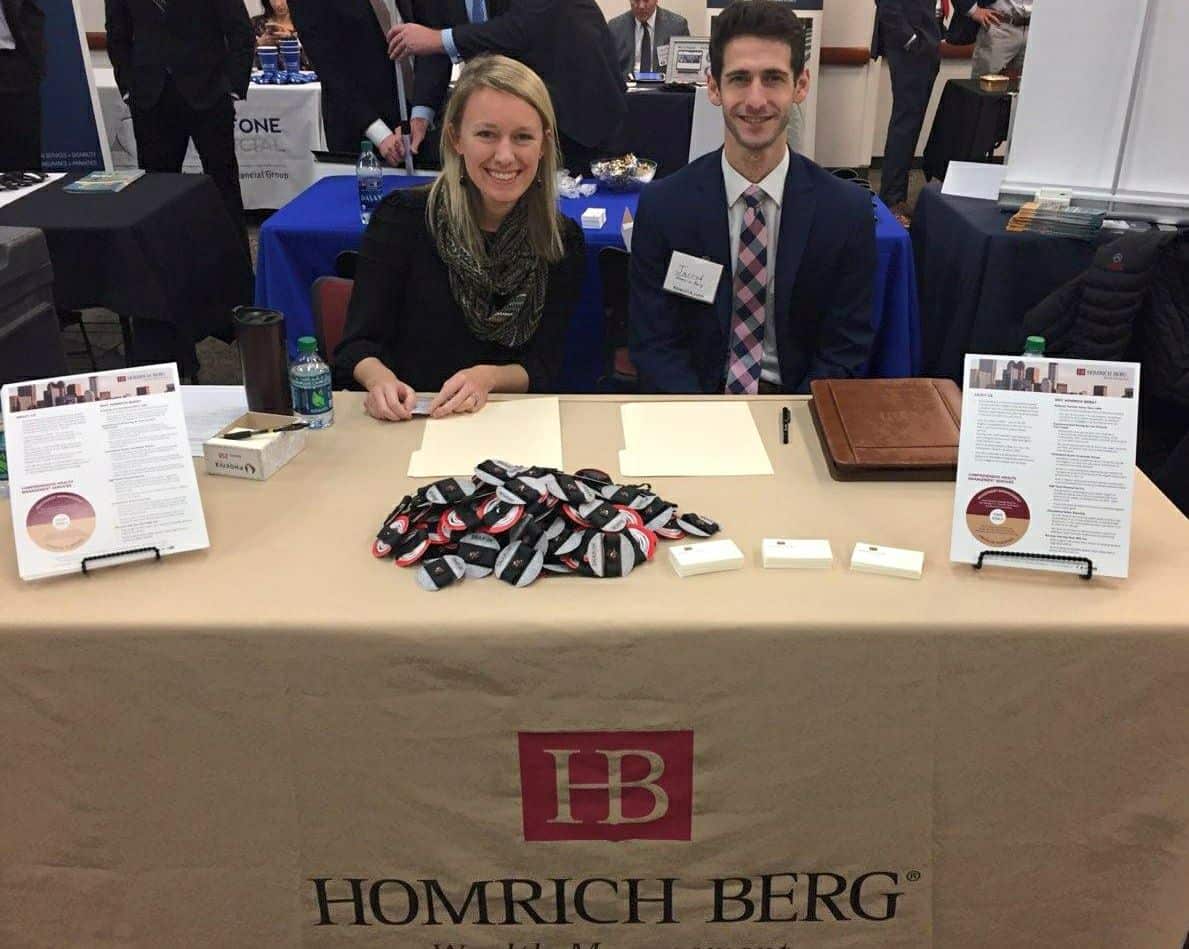 Two individuals sitting at a table with a beige cloth labeled Homrich Berg. The table has papers, a water bottle, name tags, and informational pamphlets. They are smiling and appear to be at a professional event or job fair.