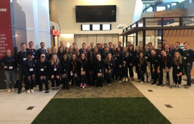 A large group of people, diverse in gender and ethnicity, posing indoors. They are smiling, wearing lanyards, and dressed casually. The background features modern office decor with glass walls and a green carpet strip on the floor.