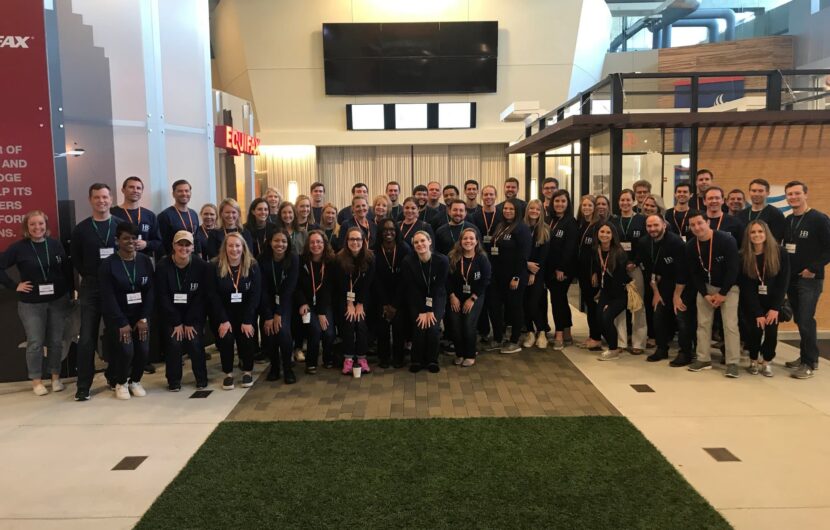 A large group of people, diverse in gender and ethnicity, posing indoors. They are smiling, wearing lanyards, and dressed casually. The background features modern office decor with glass walls and a green carpet strip on the floor.