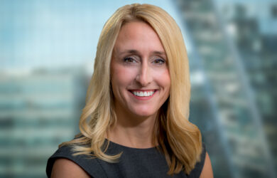 A woman with long blonde hair is smiling at the camera. She is wearing a dark sleeveless top. The background is blurred, showing indistinct tall buildings.