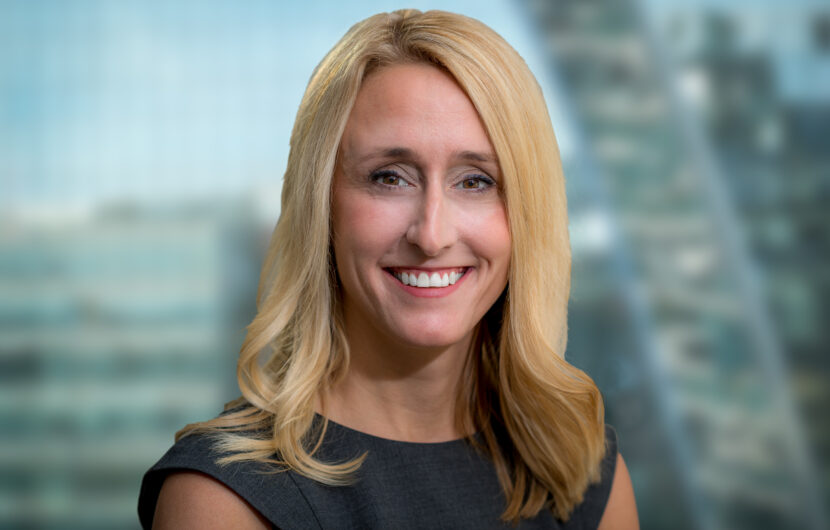 A woman with long blonde hair is smiling at the camera. She is wearing a dark sleeveless top. The background is blurred, showing indistinct tall buildings.