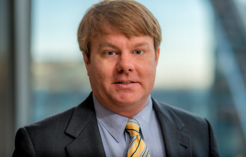 A person with short light brown hair wearing a suit, light blue shirt, and yellow striped tie is standing indoors. The background is softly blurred, suggesting a window view with natural light.