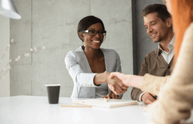 A woman in glasses smiles as she shakes hands with another individual across a table. A man sitting nearby also smiles, watching the handshake. Theres a notebook and a coffee cup on the table in a well-lit meeting room.