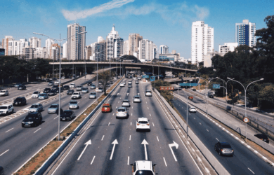 A busy multi-lane highway with cars heading in both directions. The road is flanked by trees and tall buildings under a clear blue sky. An overpass crosses the highway in the background.