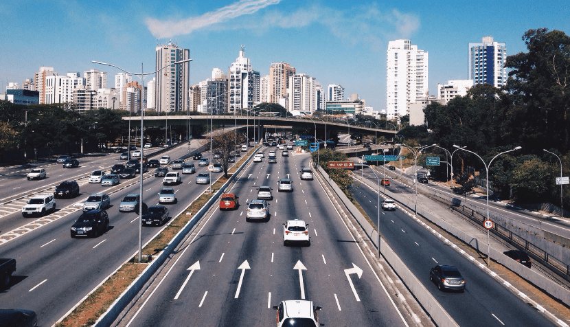 A busy multi-lane highway with cars heading in both directions. The road is flanked by trees and tall buildings under a clear blue sky. An overpass crosses the highway in the background.