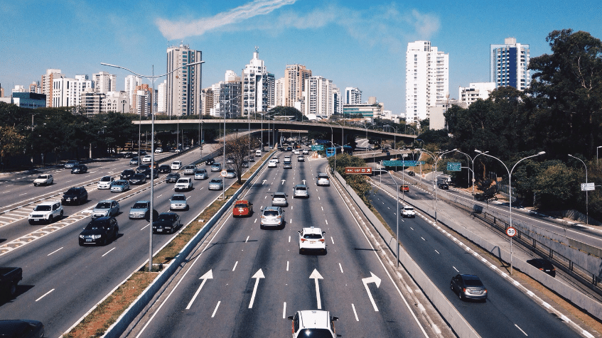 A busy multi-lane highway with cars heading in both directions. The road is flanked by trees and tall buildings under a clear blue sky. An overpass crosses the highway in the background.