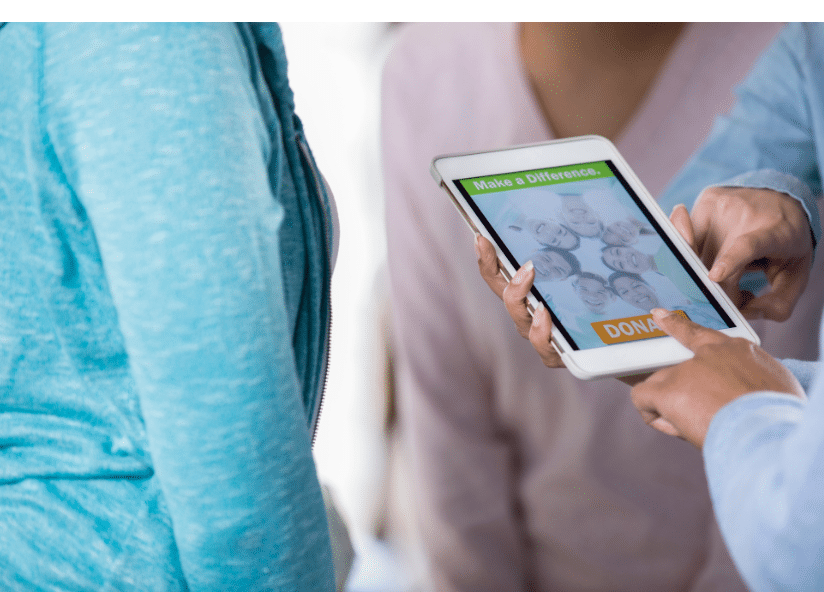 Two people holding a tablet displaying images of smiling children and a Donate button. The screen has the text Make a Difference at the top. One person points at the tablet.