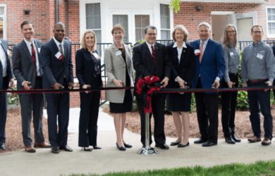 A group of people stands in front of a building, holding a long red ribbon for a ribbon-cutting ceremony. They are all dressed in formal attire.