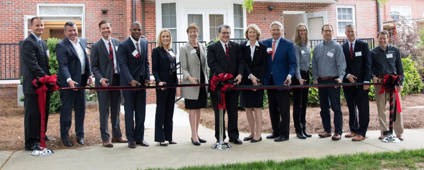 A group of people stands in front of a building, holding a long red ribbon for a ribbon-cutting ceremony. They are all dressed in formal attire.