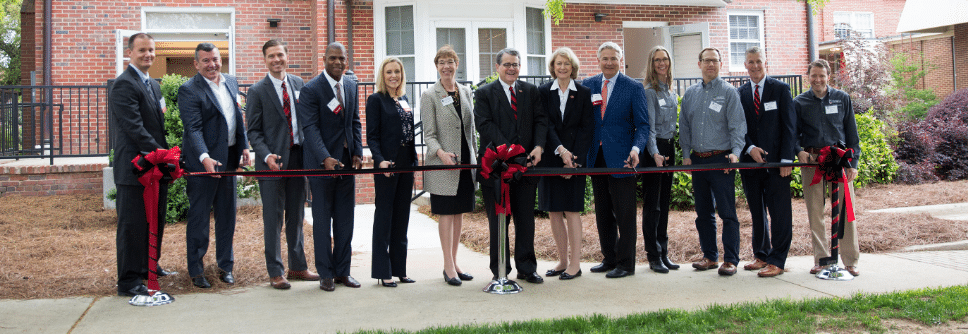 A group of people stands in front of a building, holding a long red ribbon for a ribbon-cutting ceremony. They are all dressed in formal attire.