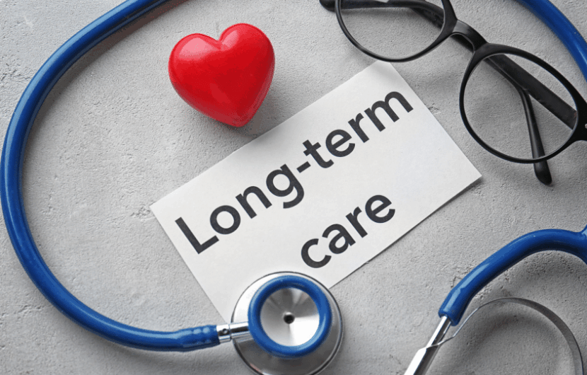 A stethoscope, red heart, and eyeglasses surround a paper with the text Long-term care on a gray surface. The items symbolize medical care and compassion.