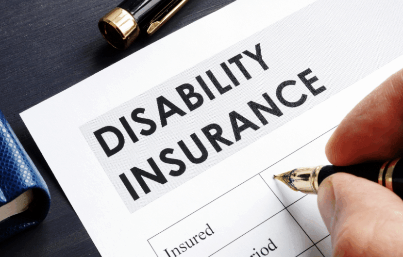 A hand holding a pen hovering over a document titled Disability Insurance. The form includes sections for Insured and Policy Period. A pen and a blue book are placed nearby on a dark surface.