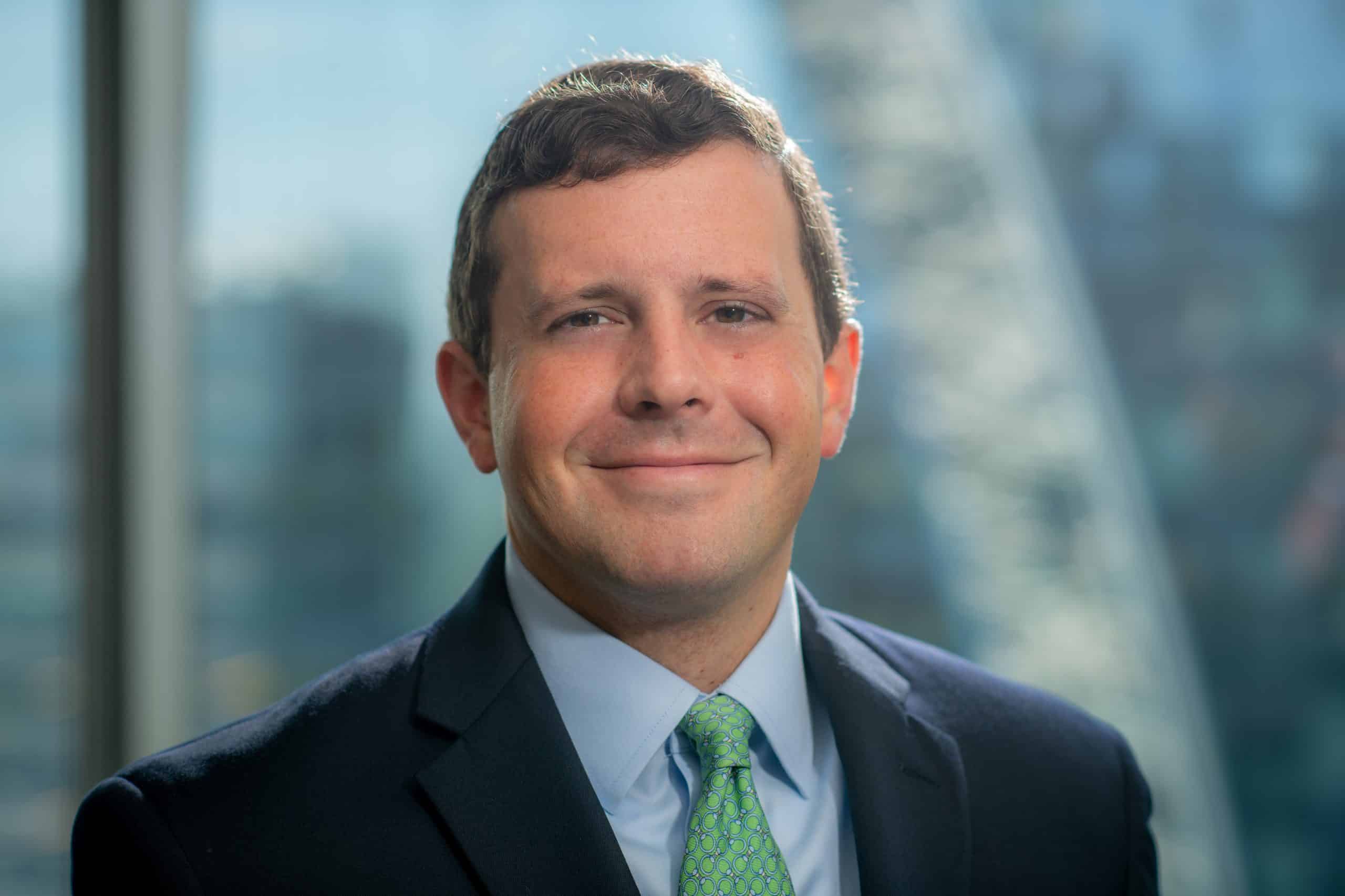 A man in a suit and green tie smiles at the camera. He has short, dark hair and the background shows a blurred cityscape with natural light coming from a nearby window.