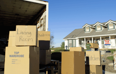 A moving truck is parked in front of a house with a For Sale sign. Several cardboard boxes labeled Living Room, Bedroom, and Kitchen are stacked outside. Two people are seen carrying boxes towards the house.