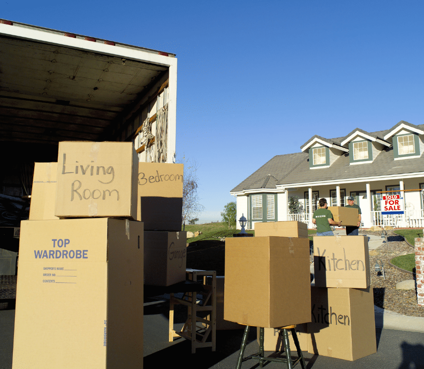 A moving truck is parked in front of a house with a For Sale sign. Several cardboard boxes labeled Living Room, Bedroom, and Kitchen are stacked outside. Two people are seen carrying boxes towards the house.