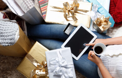 Person sitting on the floor with a tablet and a cup of coffee, surrounded by wrapped gifts with ribbons. A credit card is placed on the tablet. They are wearing jeans and a cozy sweater, next to shopping bags and presents in gold and silver wrapping.