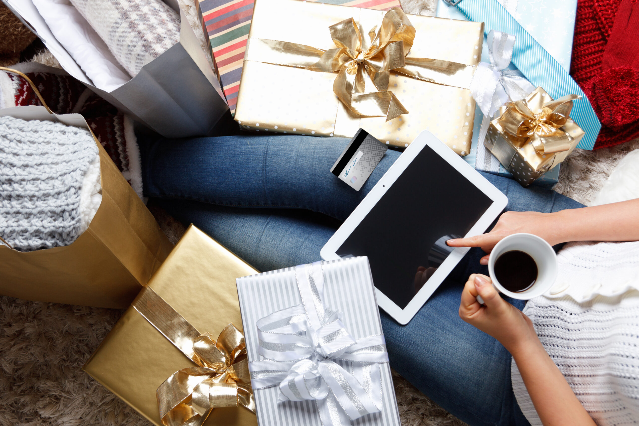 Person sitting on the floor with a tablet and a cup of coffee, surrounded by wrapped gifts with ribbons. A credit card is placed on the tablet. They are wearing jeans and a cozy sweater, next to shopping bags and presents in gold and silver wrapping.