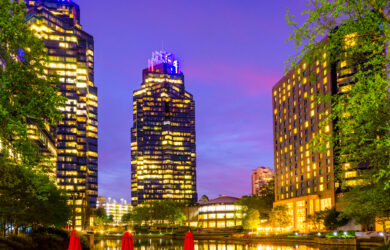 A vibrant cityscape at dusk, featuring illuminated skyscrapers reflecting on a tranquil lake. Red umbrellas and empty chairs are in the foreground, surrounded by lush greenery. The sky is a gradient of purple, pink, and blue hues.