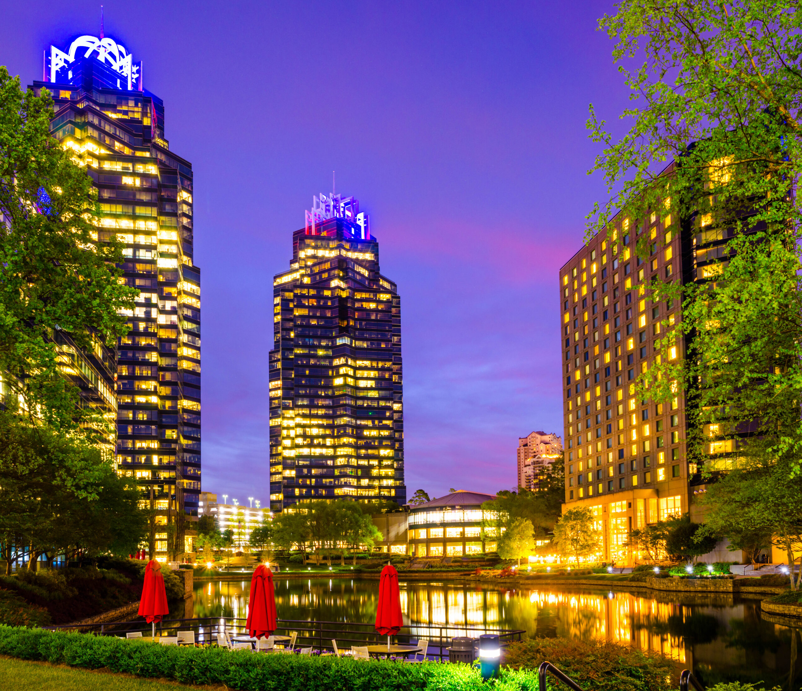 A vibrant cityscape at dusk, featuring illuminated skyscrapers reflecting on a tranquil lake. Red umbrellas and empty chairs are in the foreground, surrounded by lush greenery. The sky is a gradient of purple, pink, and blue hues.