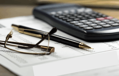 A close-up image of a calculator, a pair of glasses, and a pen placed on a paper with printed numbers. The focus is on the workspace essentials, suggesting financial or mathematical work in progress.