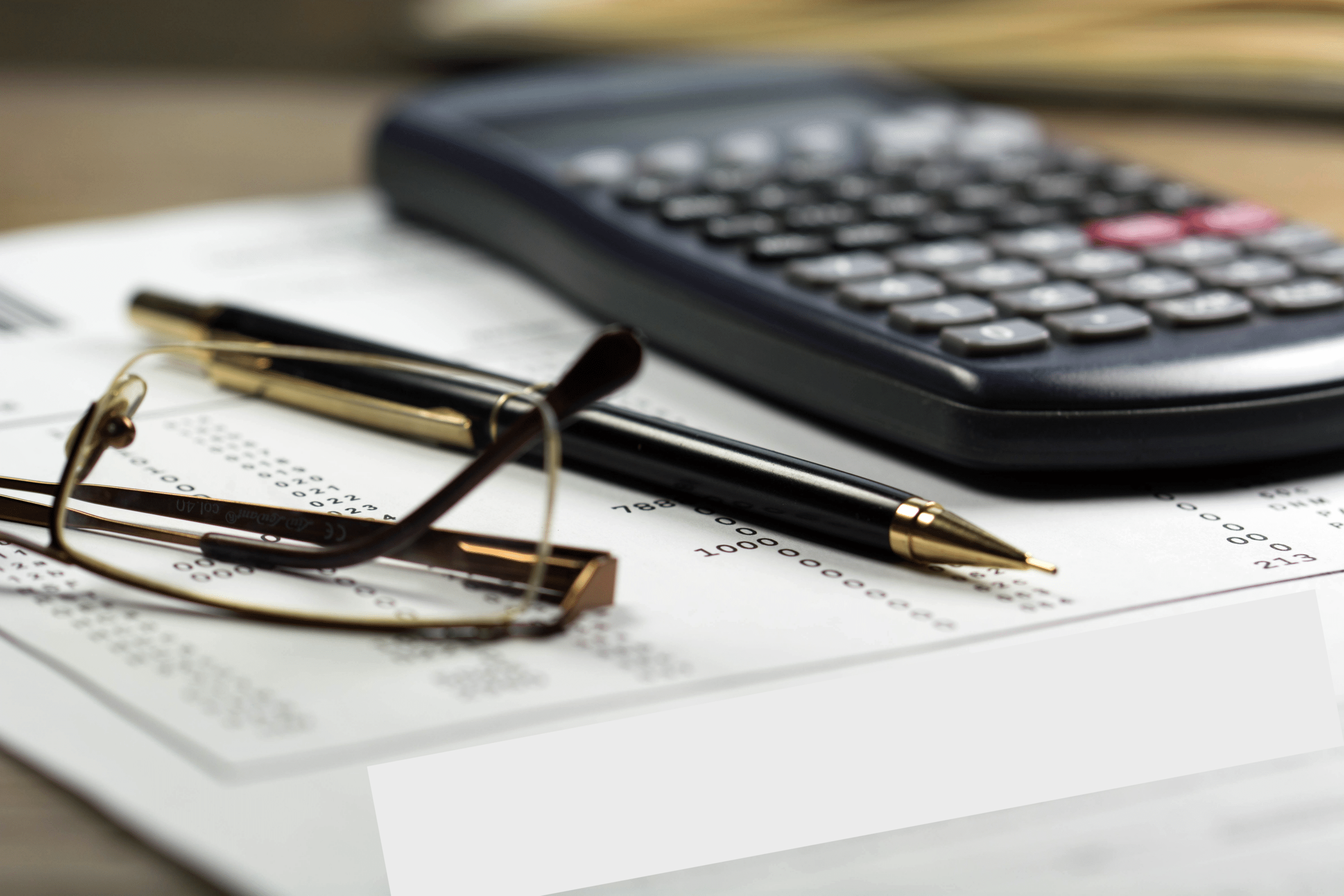 A close-up image of a calculator, a pair of glasses, and a pen placed on a paper with printed numbers. The focus is on the workspace essentials, suggesting financial or mathematical work in progress.