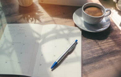 A sunlit table features an open planner displaying a monthly calendar. A blue and silver pen rests on the planner. Nearby, a steaming cup of coffee sits on a saucer, casting gentle shadows.