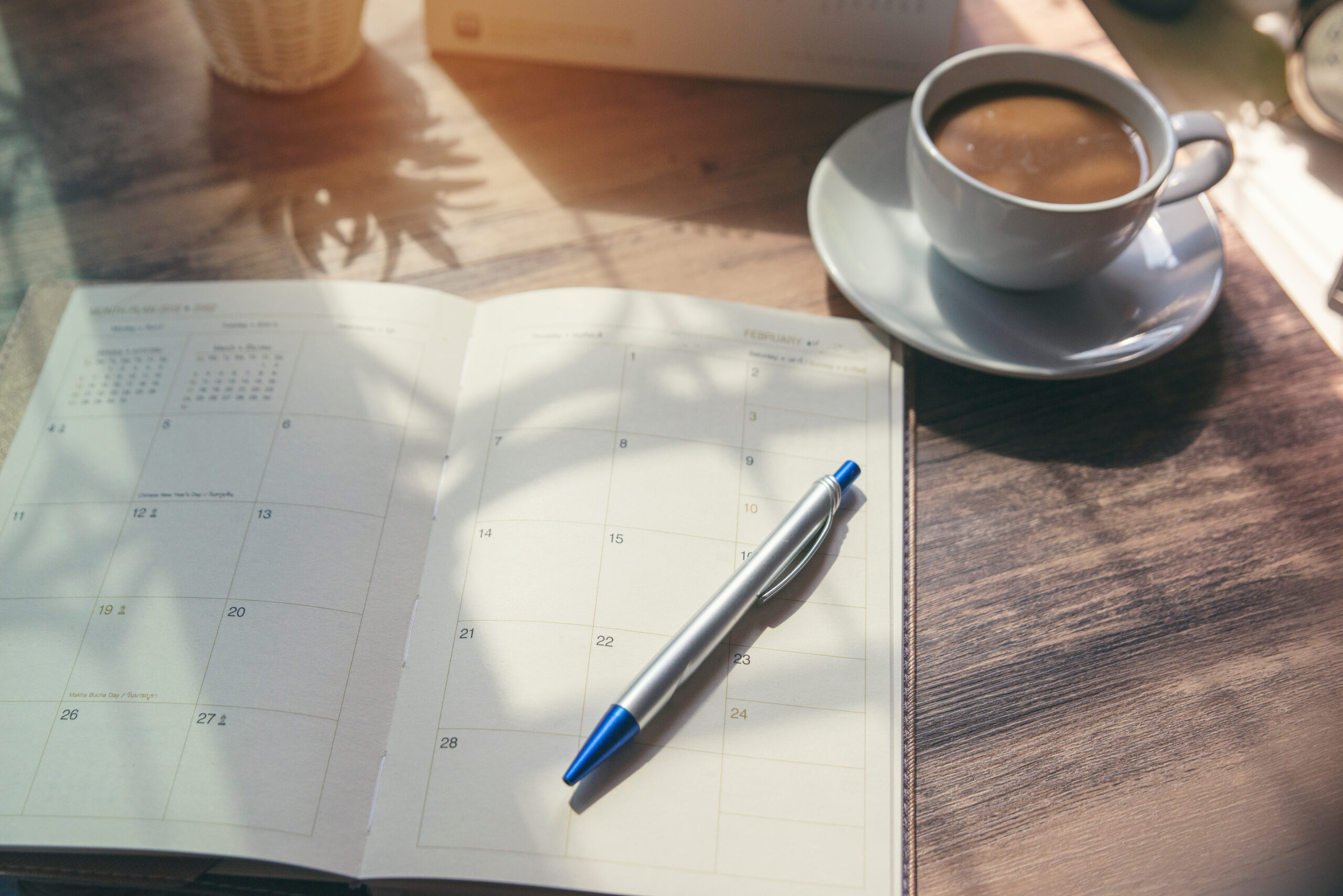A sunlit table features an open planner displaying a monthly calendar. A blue and silver pen rests on the planner. Nearby, a steaming cup of coffee sits on a saucer, casting gentle shadows.