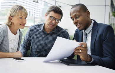 A professional meeting with three people: a woman and a man sitting beside a man in a suit who is showing them paperwork. They are at a table in a bright setting with city buildings visible outside the window.