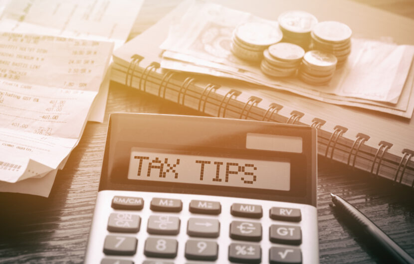 A calculator displays TAX TIPS on its screen. Nearby, there are receipts, a pen, a spiral notebook, and a stack of coins and bills. The scene is warmly lit, suggesting a focus on financial planning or tax preparation.