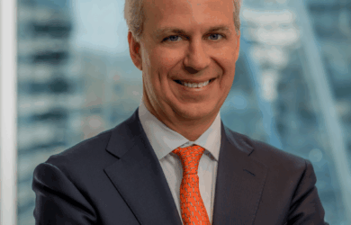 A man with short gray hair is smiling, wearing a dark suit, a white shirt, and an orange tie with blue patterns. He is standing with arms crossed in front of a window with a blurry cityscape in the background.