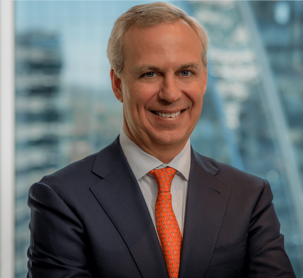 A man with short gray hair is smiling, wearing a dark suit, a white shirt, and an orange tie with blue patterns. He is standing with arms crossed in front of a window with a blurry cityscape in the background.